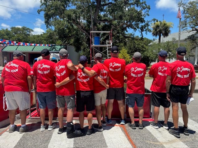 The St. James Drill Team lined up shoulder to shoulder in matching red St. James Drill Team shirts, backs to camera, facing the Bucket Brigade scaffold