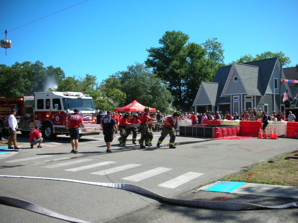 SJFD team in red shirts and turnout gear carrying hose across the competition course with a fire engine and spectators in the background