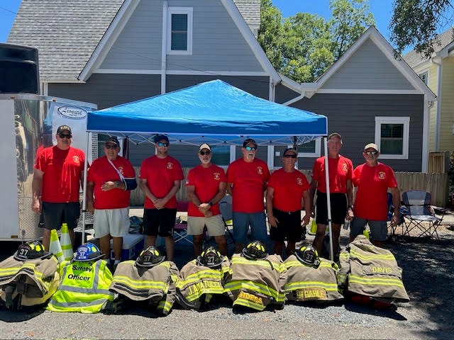 SJFD volunteers in matching red shirts pose under a blue canopy tent with turnout gear and helmets displayed in front