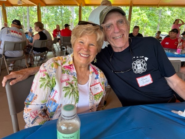Peggy and Steve Disher smile together at a picnic table with the full pavilion visible behind them