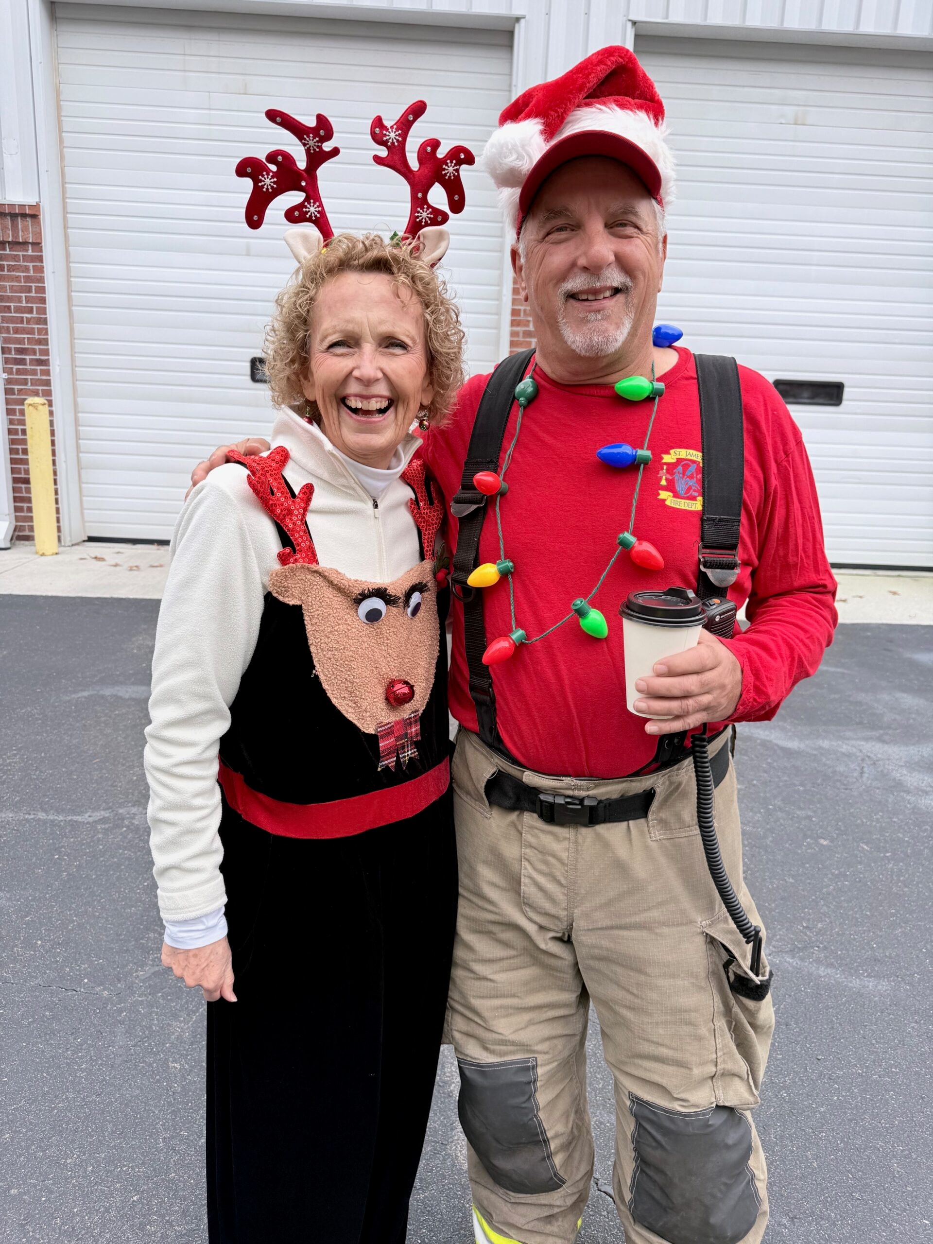 Two SJFD volunteers in festive holiday attire — reindeer antlers and Christmas lights — smiling outside the station